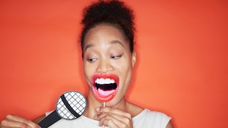 Eine Person vor orangem Hintergrund hält eine Requisite in Form eines großen Mundes vor das Gesicht und daneben ein stilisiertes Mikrofon aus Pappe. Das Motiv symbolisiert Sprache, Synchronisation und die Anpassung von Lippenbewegungen.
