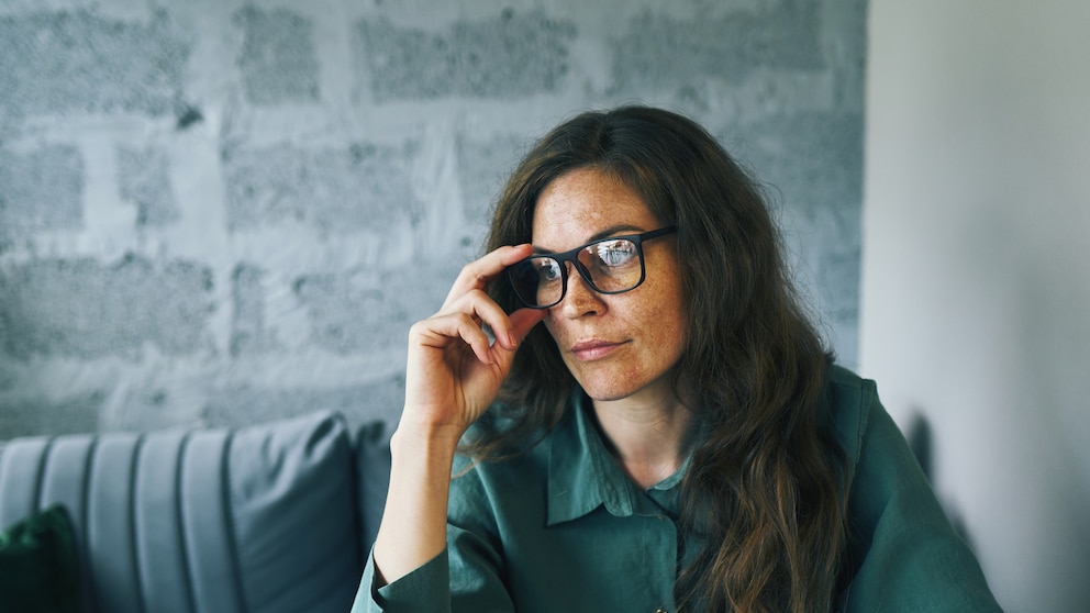 Zu sehen ist eine Person mit schwarzer Brille und langen dunklen Haaren. Eine Hand liegt am Brillengestell, während die Person an einem Tisch sitzt. Im Hintergrund befinden sich eine helle, grau strukturierte Wand sowie eine gepolsterte Sitzbank. Die Person trägt ein dunkelgrünes Oberteil. Das Bild ist im Querformat aufgenommen, wobei die Person hauptsächlich die rechte Bildhälfte einnimmt.