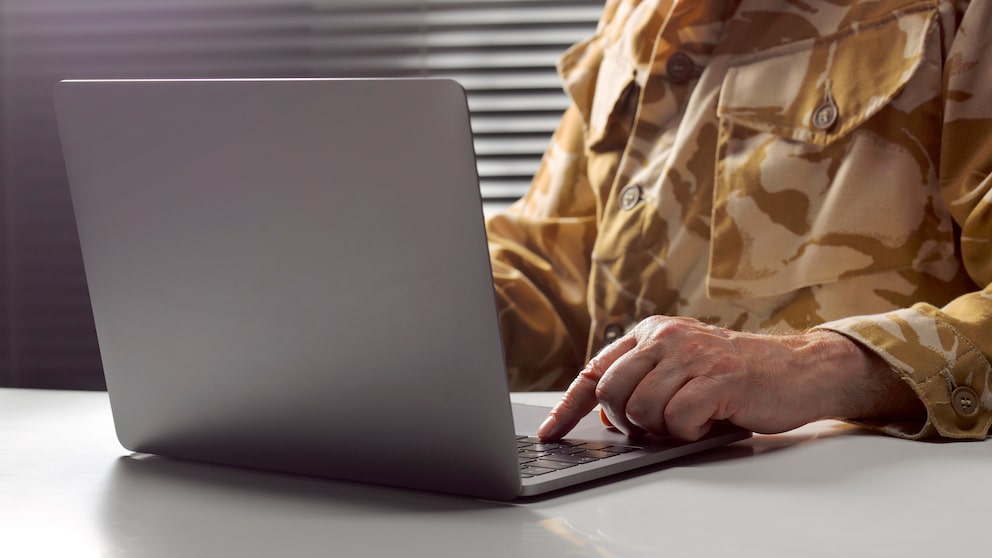 Nahaufnahme eines geöffneten Laptops auf einem Schreibtisch. Daneben sind Hände und Oberkörper einer Person in Tarnuniform zu sehen, die auf der Tastatur arbeitet. Im Hintergrund befinden sich horizontale Lamellen eines Fensters oder Rollos.