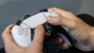 Zwei Hände halten einen weiß-schwarzen PlayStation-Controller mit Tasten und Analogsticks vor einem grauen Hintergrund