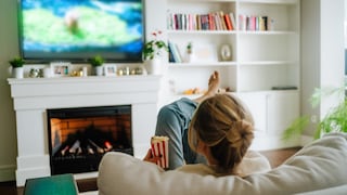 Person sitzt auf einem Sofa in einem Wohnzimmer und blickt auf einen unscharf dargestellten Fernseher. In der Hand hält die Person eine Tüte Popcorn. Vor dem Sofa befindet sich ein Kamin, daneben stehen Pflanzen und Dekorationen auf einem Regal.
