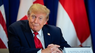 US-Präsident Donald Trump sitzt in dunklem Anzug mit roter Krawatte an einem Tisch vor mehreren Flaggen. Vor ihm steht ein Namensschild mit der Aufschrift „United States“.