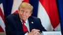 US-Pr&auml;sident Donald Trump sitzt in dunklem Anzug mit roter Krawatte an einem Tisch vor mehreren Flaggen. Vor ihm steht ein Namensschild mit der Aufschrift &bdquo;United States&ldquo;.