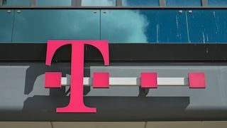 Magentafarbenes Telekom-Logo an der Fassade eines modernen Bürogebäudes. Das Logo ist vor einer Glasfront mit dunklen Fensterflächen montiert. Im oberen Bildbereich spiegeln sich Wolken in den Scheiben.