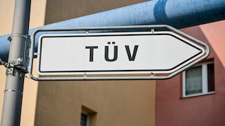 Weißes pfeilförmiges Schild mit der Aufschrift TÜV an einem Metallmast vor Gebäuden und einer blauen Rohrleitung im Hintergrund