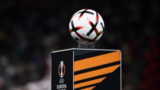Im Vordergrund ist ein weißer Fußball mit schwarzen und orangefarbenen Elementen zu sehen. Der Ball liegt auf einem schwarzen Podest mit orangefarbenem Design und dem Logo der UEFA Europa League. Der Hintergrund ist dunkel und unscharf, Personen sind dort nur schemenhaft erkennbar.