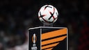 Im Vordergrund ist ein wei&szlig;er Fu&szlig;ball mit schwarzen und orangefarbenen Elementen zu sehen. Der Ball liegt auf einem schwarzen Podest mit orangefarbenem Design und dem Logo der UEFA Europa League. Der Hintergrund ist dunkel und unscharf, Personen sind dort nur schemenhaft erkennbar.