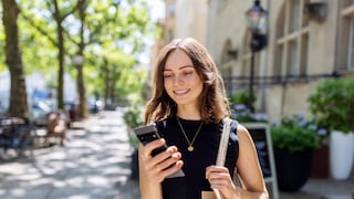 Person mit schulterlangem Haar steht auf einem sonnigen Gehweg zwischen Bäumen und Gebäuden und blickt auf ein Smartphone in der Hand. Über einer Schulter hängt eine helle Stofftasche, im Hintergrund sind unscharf eine Straße, Fahrräder und Hausfassaden zu sehen. Geben Sie Ihr Feedback zu BizChat ab.
