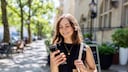 Person mit schulterlangem Haar steht auf einem sonnigen Gehweg zwischen B&auml;umen und Geb&auml;uden und blickt auf ein Smartphone in der Hand. &Uuml;ber einer Schulter h&auml;ngt eine helle Stofftasche, im Hintergrund sind unscharf eine Stra&szlig;e, Fahrr&auml;der und Hausfassaden zu sehen. Geben Sie Ihr Feedback zu BizChat ab.
