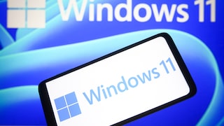 Ein Smartphone zeigt das Windows-11-Logo vor einem Bildschirm mit großem Windows-11-Schriftzug und blauem Hintergrund