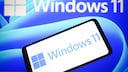 Ein Smartphone zeigt das Windows-11-Logo vor einem Bildschirm mit gro&szlig;em Windows-11-Schriftzug und blauem Hintergrund