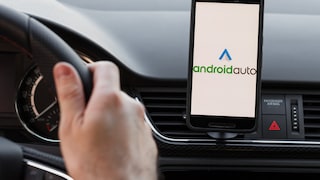 Eine Person sitzt im Auto mit den Händen am Lenkrad. Man sieht ein Handy in einer Halterung am Armaturenbrett auf dem Android Auto geöffnet ist.
