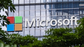Microsoft-Logo und weißer Microsoft-Schriftzug an der Glasfassade eines Bürogebäudes. Im Vordergrund sind unscharfe grüne Blätter und Äste zu sehen, die den unteren und seitlichen Bildbereich teilweise verdecken.
