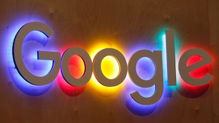 Großes Google-Logo aus weißen dreidimensionalen Buchstaben an einer Holzwand. Hinter den einzelnen Buchstaben leuchten farbige Lichtflächen in Blau, Rot, Gelb und Grün, die auf die Wand abstrahlen. Das Foto zeigt das Logo frontal und füllt den Großteil des Bildes aus.