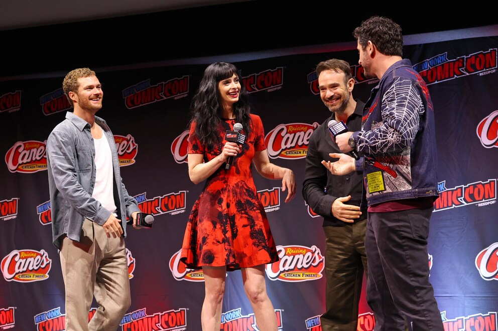 Four people stand on a stage in front of a backdrop with the logos of New York Comic Con and Raising Cane’s. Several people hold microphones and talk to each other. One person wears a red-black dress, the others dark or light casual clothing. The stage is lit by spotlights, and the event logos are repeatedly visible in the background.