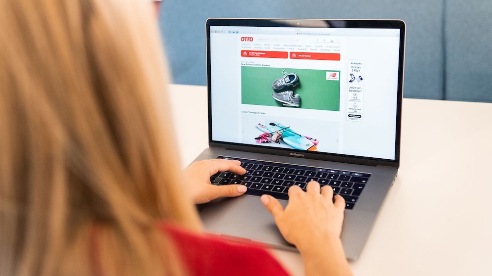 Zu sehen ist eine Person, die an einem MacBook Pro sitzt und die Webseite des Onlinehändlers Otto geöffnet hat. Auf dem Bildschirm sind verschiedene Produkte und Angebote des Otto-Onlineshops zu sehen. Die Person befindet sich im Vordergrund und bedient das Notebook mit beiden Händen. Im Hintergrund ist eine schlichte blaue Wand zu erkennen.