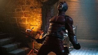 Person in einem dunkelroten Superheldenkostüm mit Maske steht in einem gemauerten Raum unter einem Rundbogen. In einer Hand hält die Figur einen Schlagstock. Der Raum wird von warmem orangefarbenem Licht auf der linken und kühlerem Licht auf der rechten Seite beleuchtet. Im Hintergrund sind eine Treppe, Baustrahler und weitere technische Geräte zu sehen.