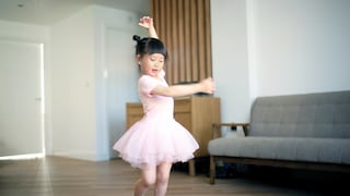 Ein junges Mädchen mit dunklen, hochgebundenen Haaren tanzt in einem hellen Wohnzimmer. Es trägt ein rosafarbenes Ballettkleid mit Tüllrock und passende Ballettschuhe. Ein Arm ist über den Kopf gehoben, der andere seitlich ausgestreckt. Im Hintergrund stehen ein graues Sofa und eine Holzkommode.