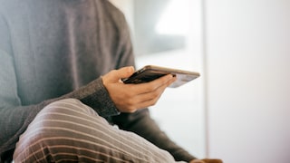 Person sitzt in einem hellen Raum und hält ein Smartphone waagerecht in einer Hand. Die Person trägt ein dunkelgraues Oberteil und eine grau gestreifte Hose. Der Blick ist auf das Smartphone gerichtet, der Hintergrund ist hell und unscharf.