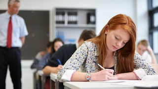 Eine rothaarige Schülerin sitzt an einem Tisch im Klassenzimmer und schreibt mit einem Stift auf Papier. Im Hintergrund sitzen weitere Schüler an ihren Tischen, während ein Lehrer mit roter Krawatte im Raum steht.