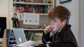 Kind sitzt an einem Schreibtisch vor einem geöffneten Laptop, surft im Internet und trägt weiße kabelgebundene Ohrhörer. Eine Hand liegt am Kinn, die andere hält einen Stift. Auf dem Tisch liegen mehrere Blätter, Bücher und schwarze Kopfhörer. Im Hintergrund steht ein Regal mit Büchern und weiteren Gegenständen.