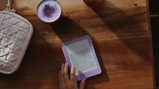 Eine Hand berührt einen violetten Amazon-Kindle-E-Reader auf einem dunklen Holztisch. Auf dem Display ist eine Seite mit deutschsprachigem Fließtext zu sehen. Neben dem E-Reader stehen eine Tasse mit einem hellvioletten Getränk und Schaum sowie eine helle geflochtene Handtasche. Auf dem Tisch fallen Sonnenlicht und Schattenmuster. Die Fingernägel der Hand sind passend zum E-Reader violett lackiert.