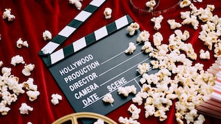 Eine Filmklappe mit der Aufschrift „Hollywood Production“ liegt auf einem roten Stoffhintergrund. Um die Filmklappe herum sind zahlreiche Popcornstücke verteilt. Im unteren Bildbereich ist ein Teil einer Filmrolle zu sehen, im oberen Bereich ein Streifen Film. Rechts befindet sich ein rot-weiß gestreifter Popcornbehälter. Die Szene ist von oben fotografiert und zeigt verschiedene Kino- und Filmrequisiten.