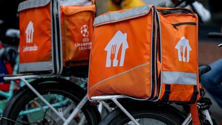Mehrere orangefarbene Lieferando-Liefertaschen mit weißen Logos und grauen Reflektorstreifen stehen auf den Gepäckträgern von Fahrrädern. Auf einer der Taschen ist zusätzlich das Logo der UEFA Champions League zu sehen. Im unscharfen Hintergrund befinden sich mehrere Personen, darunter eine Person in einer gelben Jacke.