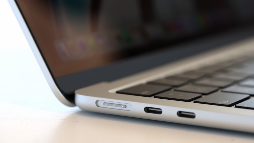Nahaufnahme der linken Seite eines silberfarbenen Notebooks. Sichtbar sind die Tastatur, der geöffnete Bildschirmrand sowie zwei USB-C-Anschlüsse an der Gehäuseseite. Daneben befindet sich ein magnetischer Ladeanschluss mit fünf Kontaktpunkten. Der Hintergrund ist unscharf, das Gerät steht auf einer hellen Oberfläche.