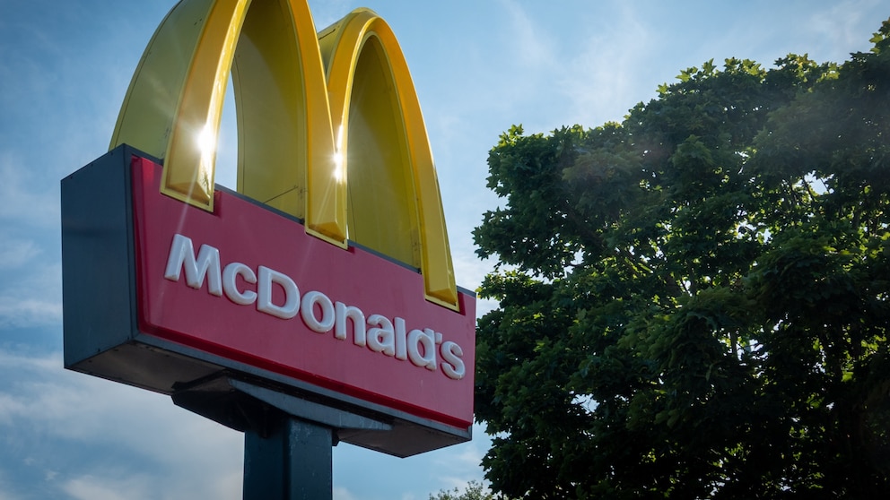 Nahaufnahme eines großen McDonald's-Werbeschilds vor blauem Himmel. Auf dem roten Schild steht der weiße Schriftzug „McDonald's“, darüber befinden sich die beiden gelben Bögen des Firmenlogos. Sonnenlicht scheint durch die Bögen. Rechts im Bild ist die grüne Krone eines Baumes zu sehen.