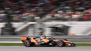 Orange-schwarzer Formel-1-Rennwagen von McLaren auf einer Rennstrecke in Seitenansicht. Das Fahrzeug fährt auf grauem Asphalt und trägt mehrere Sponsorenlogos auf Karosserie und Heckflügel. Die Reifen besitzen farbige Markierungen an den Felgen. Im Hintergrund sind Tribünen mit zahlreichen Zuschauern zu sehen, die durch die Bewegung des Fahrzeugs und eine lange Belichtungszeit unscharf erscheinen. Vor den Tribünen verlaufen Leitplanken und Streckenbegrenzungen.