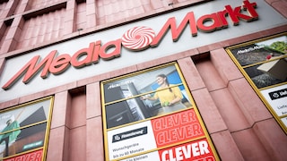 Fassade eines MediaMarkt-Geschäfts in Untersicht. Im oberen Bereich befindet sich ein großes „MediaMarkt“-Logo mit rotem Schriftzug und rot-weißem Spiralsymbol. Darunter sind mehrere große Schaufenster mit Werbeplakaten zu sehen. Auf einem Plakat steht mehrfach das Wort „CLEVER“, auf einem anderen werden Finanzierungsmöglichkeiten für Wunschgeräte beworben. Die Fassade ist in rosa- und rotfarbenen Tönen gestaltet.