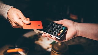 Nahaufnahme einer kontaktlosen Kartenzahlung. Eine Hand hält eine orangefarbene Karte über ein schwarzes Kartenlesegerät, das von einer zweiten Hand gehalten wird. Das Terminal verfügt über ein Ziffernfeld sowie grüne, gelbe und rote Funktionstasten. Der Hintergrund ist dunkel und unscharf.