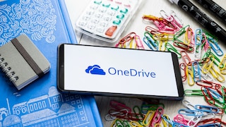 Smartphone mit dem OneDrive-Logo auf dem Display liegt auf einem Schreibtisch. Um das Gerät herum befinden sich ein blaues Notizbuch, ein kleiner Spiralblock, ein Taschenrechner, mehrere Filzstifte sowie zahlreiche bunte Büroklammern in Rot, Gelb, Grün, Blau und Weiß. Der Bildschirm zeigt auf weißem Hintergrund das OneDrive-Logo und den Schriftzug „OneDrive“.