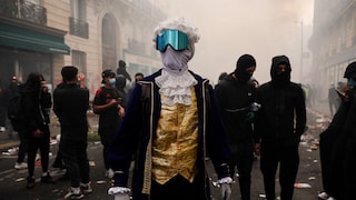 Eine Person in historischer Kleidung mit weißer Lockenperücke, goldener Weste und dunkelblauem Mantel steht mitten auf einer Straße. Das Gesicht ist mit einem grauen Tuch und einer großen, blau verspiegelten Schutzbrille verdeckt. Im Hintergrund befinden sich zahlreiche Menschen, teilweise mit schwarzen Gesichtsmasken. Die Straße ist mit Müll übersät und von dichtem Rauch oder Nebel erfüllt.