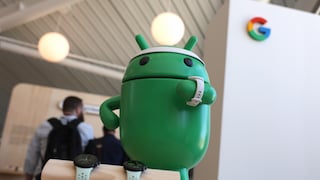 Große grüne Android-Figur steht auf einem Messestand und trägt eine weiße Smartwatch am Arm. Vor der Figur liegen zwei weitere Smartwatches mit hellen Armbändern auf einer Holzhalterung. Rechts im Hintergrund ist das mehrfarbige Google-Logo zu sehen, links mehrere unscharf abgebildete Personen.