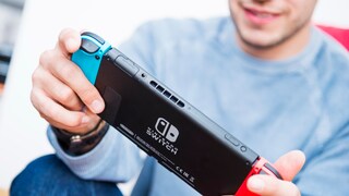 Ein Mann in einem hellblauen Pullover hält eine Nintendo Switch mit beiden Händen im Querformat. Zu sehen ist die schwarze Rückseite der Konsole mit dem weißen Nintendo-Switch-Logo. Links ist ein blauer Joy-Con-Controller angebracht, rechts ein roter. Der Mann blickt lächelnd auf die Konsole.