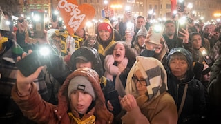 Eine große Menschenmenge steht nachts auf einem beleuchteten Platz vor historischen Gebäuden. Viele Personen halten Smartphones mit eingeschalteten Taschenlampen hoch. In der Bildmitte ist ein orangefarbenes Herz mit der weißen Aufschrift „OSTRAVA“ zu sehen. Mehrere Menschen tragen Regenjacken, Mützen und Kapuzen. Im Hintergrund wird eine tschechische Flagge hochgehalten.