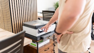 Person steht vor einem Drucker in einem Büro und berührt mit einem Finger das beleuchtete Display des Geräts. Der Drucker steht auf einem kleinen Schrank mit Schubladen. Im Hintergrund sind Holzwandpaneele, Schreibtische und Bürostühle zu sehen. Die Aufnahme zeigt hauptsächlich den Drucker, den Arm der Person und den Bedienbereich des Geräts.