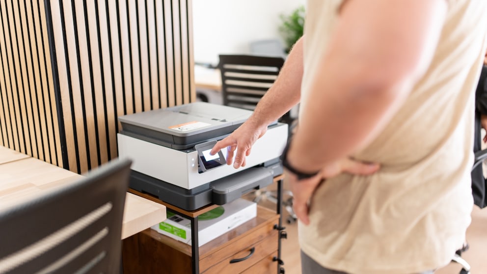 Person steht vor einem Drucker in einem Büro und berührt mit einem Finger das beleuchtete Display des Geräts. Der Drucker steht auf einem kleinen Schrank mit Schubladen. Im Hintergrund sind Holzwandpaneele, Schreibtische und Bürostühle zu sehen. Die Aufnahme zeigt hauptsächlich den Drucker, den Arm der Person und den Bedienbereich des Geräts.