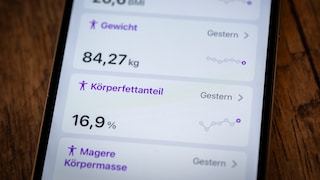 Nahaufnahme eines Smartphones auf einer Holzoberfläche. Auf dem Display werden Gesundheitsdaten angezeigt, darunter ein BMI von 26,6, ein Gewicht von 84,27 Kilogramm und ein Körperfettanteil von 16,9 Prozent. Neben den Werten sind kleine Verlaufsgrafiken zu sehen.