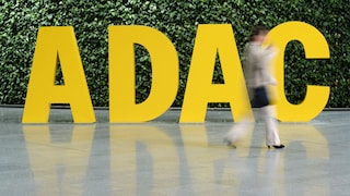 Das Bild zeigt große gelbe Buchstaben mit dem Schriftzug „ADAC“ vor einer dichten grünen Pflanzenwand. Im Vordergrund läuft eine Person durch das Bild und ist durch die Bewegung deutlich unscharf. Der Schriftzug sowie Teile der Person spiegeln sich auf dem glatten Boden.