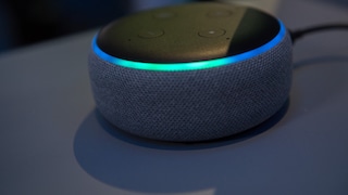 Zu sehen ist ein kompakter, runder Smart Speaker mit dunkelgrauem Stoffbezug und schwarzer Oberseite. Am oberen Rand leuchtet ein blau-türkiser Lichtring. Auf der Oberseite sind mehrere Bedientasten zu erkennen. Der Lautsprecher steht auf einer dunklen Oberfläche, während der Hintergrund unscharf und ebenfalls dunkel gehalten ist.