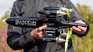 Eine Person hält eine schwarze Drohne mit mehreren Propellern in den Händen. Auf dem Rumpf stehen unter anderem Quantum Systems und N3XT, im Hintergrund ist Vegetation zu sehen