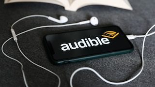 Smartphone mit Audible-Logo liegt neben weißen Kopfhörern und einem aufgeschlagenen Buch