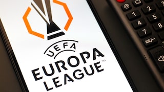 Auf dem Foto liegt ein Smartphone neben einer schwarzen TV-Fernbedienung. Auf dem hellen Display des Smartphones ist groß das schwarz-orange Logo der UEFA Europa League zu sehen. Rechts daneben sind unter anderem der rote Ein-Aus-Schalter und die Zifferntasten der Fernbedienung sichtbar.
