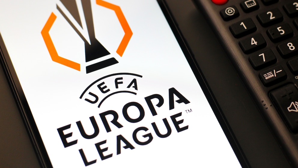Auf dem Foto liegt ein Smartphone neben einer schwarzen TV-Fernbedienung. Auf dem hellen Display des Smartphones ist groß das schwarz-orange Logo der UEFA Europa League zu sehen. Rechts daneben sind unter anderem der rote Ein-Aus-Schalter und die Zifferntasten der Fernbedienung sichtbar.