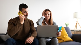 Zwei Personen sitzen vor einem Laptop auf einem Sofa. Eine Person telefoniert mit konzentriertem Gesichtsausdruck, die andere blickt mit gerunzelter Stirn auf den Bildschirm und hält eine Bankkarte in der Hand
