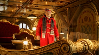 Auf dem Bild steht eine Person in einem aufwendig gestalteten, weihnachtlich wirkenden Schlitten. Die Person trägt einen langen roten Mantel, eine rot gemusterte Mütze und einen schwarz-rot-weißen Schal. Der Schlitten ist mit goldfarbenen Verzierungen, dunklen Flächen und rotem Samt ausgestattet. Mehrere leuchtende Laternen sorgen für warmes Licht. Im Hintergrund sind verzierte Wände, Bögen und eine spiegelnde Decke zu sehen.