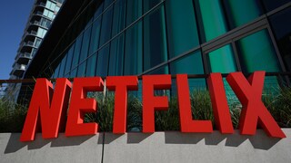 Roter Netflix-Schriftzug vor der Glasfassade eines modernen Gebäudes unter blauem Himmel.